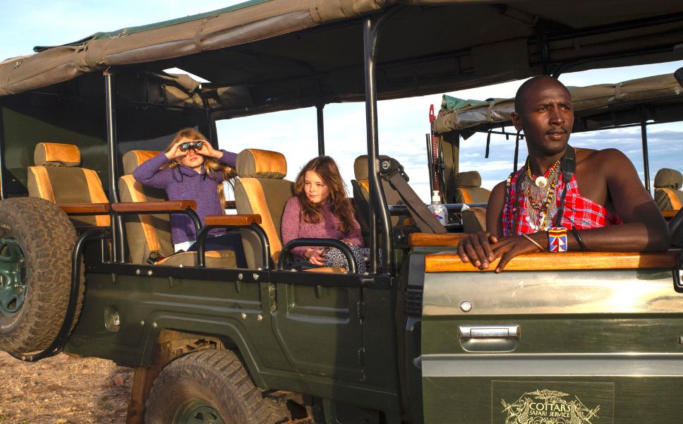 Two young guests in a safari game vehicle search for wildlife through binoculars, with the help of a local Maasai Guide.