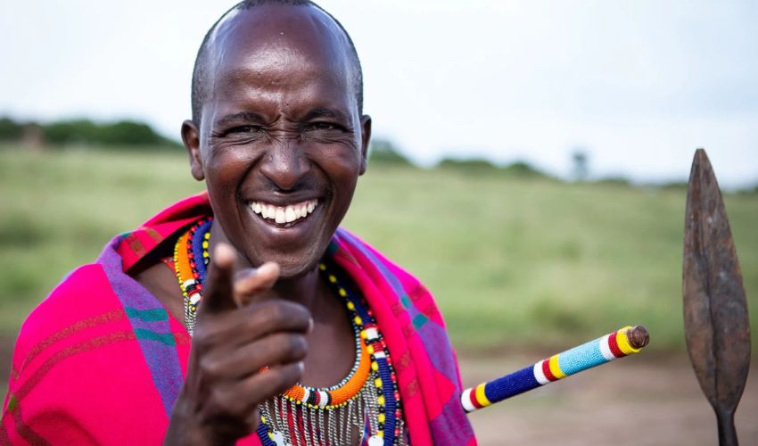A joyful Maasai man in traditional beadwork and a bright shuka, smiling and pointing at the camera, with a spear in hand against the backdrop of the Kenyan savannah.