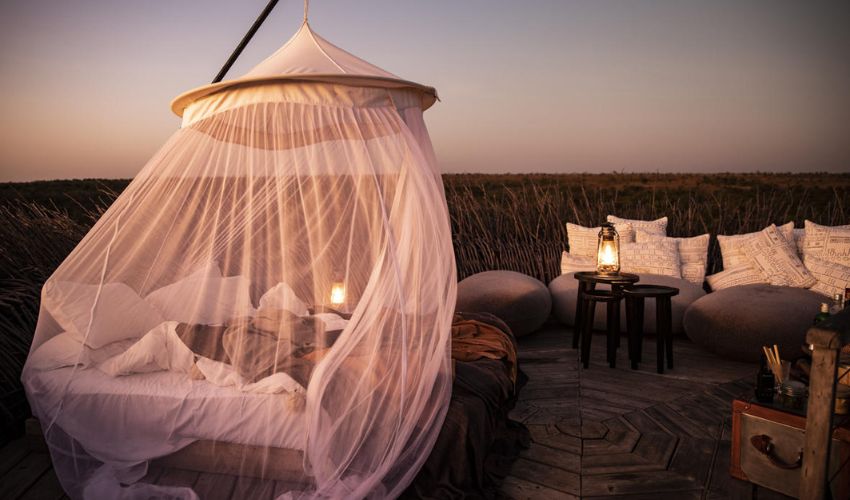 A dreamy outdoor bed draped in a white canopy netting is softly lit by lanterns at sunset, surrounded by cozy seating in the African wilderness.
