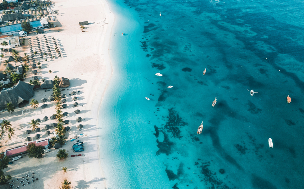 Aerial view of a white sand beach in Zanzibar with turquoise waters, scattered boats, and rows of beach umbrellas and palm trees.